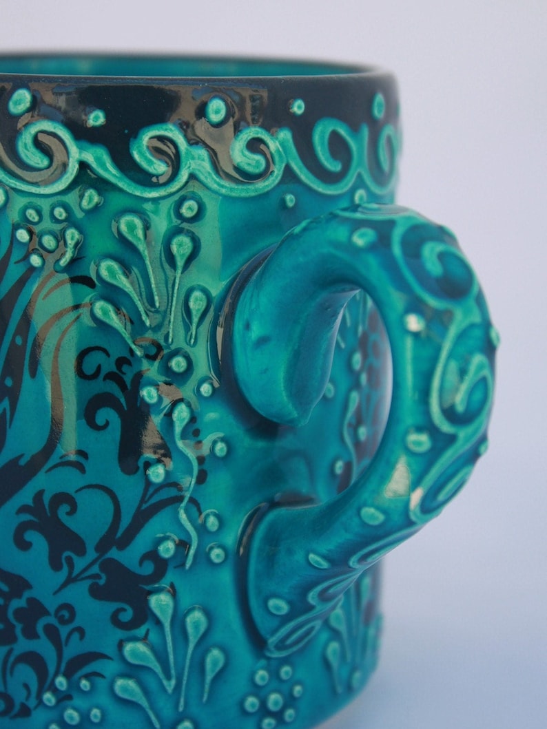 Puede incluir: Una taza de cer&aacute;mica azul turquesa con un dise&ntilde;o floral negro y un asa &uacute;nica. El asa tiene forma de cuerda trenzada y presenta un dise&ntilde;o en relieve.