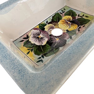 May include: A white ceramic sink with a blue rim and a floral design featuring purple, yellow, and green pansies.