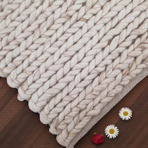 May include: A chunky knit blanket in cream and beige tones, with a scalloped edge. The thick yarn is arranged in a braided pattern. Two daisies and a red rose petal are placed on a dark wood surface.