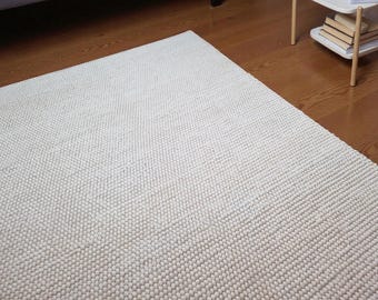 Pode incluir: Tapete texturizado branco sujo com uma pilha densa e em laço. O tapete é retangular e cobre uma parte de um chão de madeira. A superfície do tapete é composta por pequenos laços espaçados uniformemente, criando uma aparência macia e tátil.