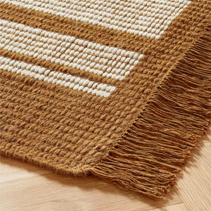 May include: A close-up of a woven rug with a textured surface. The rug features alternating stripes of tan and off-white. The edge of the rug has a fringe detail. The rug is on a wooden floor.