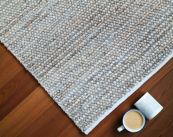 May include: A textured, rectangular rug with a mix of light blue and tan fibers. The rug is placed on a wooden floor, with a cup of coffee and a book nearby. The rug's design is a neutral color palette.
