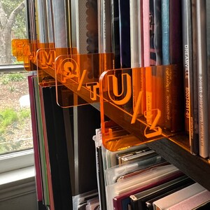 May include: Orange acrylic record dividers with letters and numbers, organizing vinyl records on a shelf. The dividers spell out "M Q T U 1 2" and are placed among various album covers. A window is visible in the background.