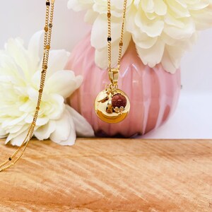 May include: A gold chain necklace with a round pendant featuring a small red stone and a gold leaf design. The necklace is displayed against a pink background with white flowers.