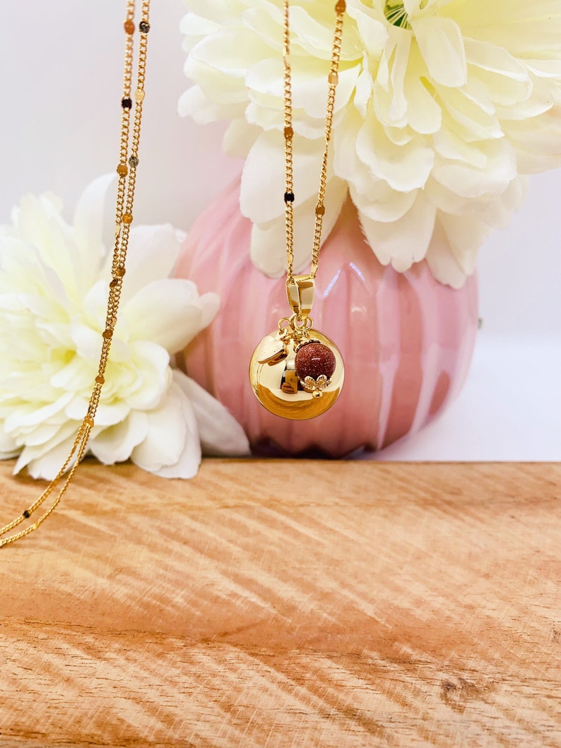 May include: A gold chain necklace with a round pendant featuring a small red stone and a gold leaf design. The necklace is displayed against a pink background with white flowers.
