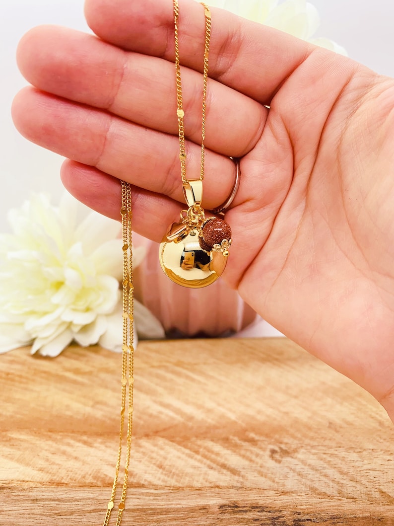 May include: A gold chain necklace with a round gold pendant and a small brown stone charm. The necklace is on a hand.