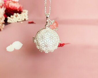 May include: A silver necklace with a delicate, white, filigree ball charm and a pink crystal bead. The necklace is hanging against a pink background.