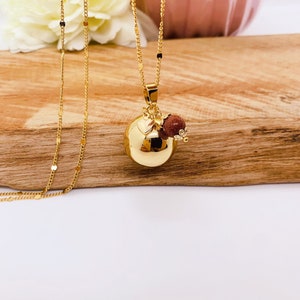May include: A gold chain necklace with a round gold pendant and a small red stone charm. The necklace is on a wooden surface.