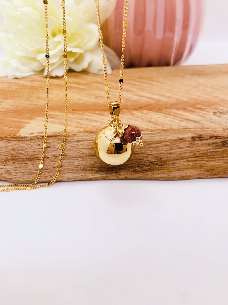 May include: A gold chain necklace with a round gold pendant and a small red stone charm. The necklace is on a wooden surface.