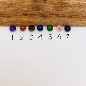 May include: Seven round beads in different colors, numbered 1 through 7. The beads are purple, brown, tiger's eye, blue, green, pink, and dark blue.