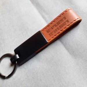 Puede incluir: Un llavero de cuero marrón con un acento de metal negro y un llavero plateado. La correa de cuero está cosida y presenta coordenadas grabadas: 40°47'6.3276"N 73°58'5.8260"W.