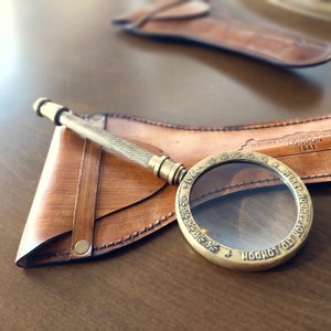 May include: A vintage brass magnifying glass with a textured handle rests on a brown leather case. The magnifying glass has a circular lens and is engraved with the text "HENRY HUGHES & SON LTD LONDON 1941".