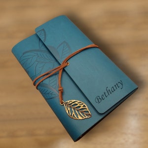 Può includere: Un diario in pelle blu con un cinturino in pelle marrone e un ciondolo a forma di foglia d'oro. Il diario ha un design floreale e il nome "Bethany" è inciso sulla parte anteriore.