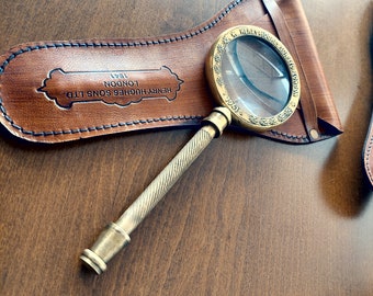 Puede incluir: Una lupa de latón con una funda de cuero. La funda es marrón y tiene el texto "HENRY HUGHES SONS LTD LONDON 1861" grabado en relieve.