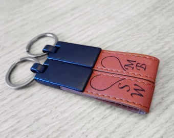 Könnte beinhalten: Ein brauner Leder-Schlüsselanhänger mit zwei Metallschlüsselringen und zwei blauen Metallplatten. Das Leder ist mit zwei ineinander verschlungenen Herzen und den Initialen "M.S" und "M.B" graviert.