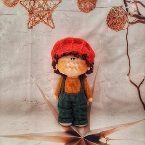Könnte beinhalten: Eine gehäkelte Puppe mit einem roten Hut, braunen Haaren und einem grün-orangenen Outfit. Die Puppe steht auf einer weißen Oberfläche mit einem braunen sternförmigen Design.
