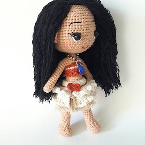Puede incluir: Mu&ntilde;eca de crochet con cabello negro largo, vistiendo un top marr&oacute;n y naranja, una falda blanca con un lazo rojo y un collar azul con un colgante de concha.