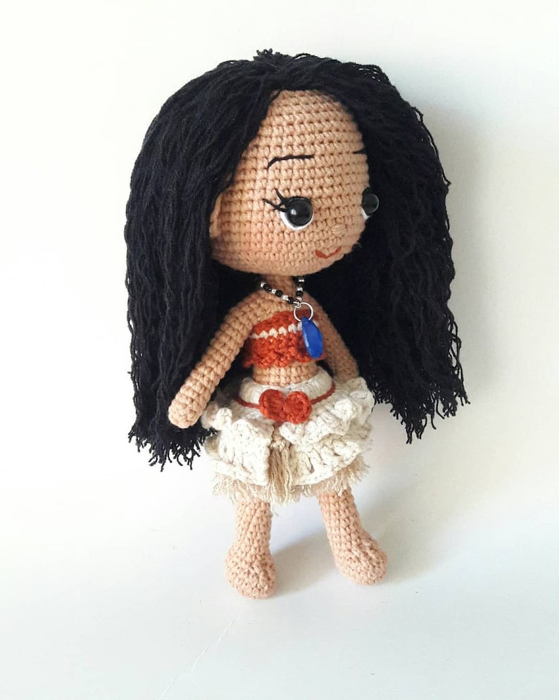 Puede incluir: Mu&ntilde;eca de crochet con cabello negro largo, vistiendo un top marr&oacute;n y naranja, una falda blanca con un lazo rojo y un collar azul con un colgante de concha.