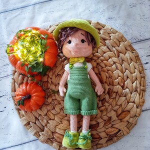 Puede incluir: Una muñeca de crochet con un sombrero verde, un mono verde y zapatos verdes. La muñeca está sentada en una estera de paja tejida con dos calabazas naranjas.