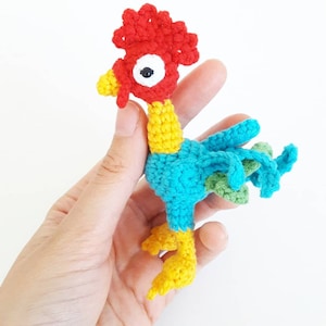 Puede incluir: Un juguete de gallo de crochet con cresta roja, pico amarillo, cuerpo azul y plumas de cola verdes.