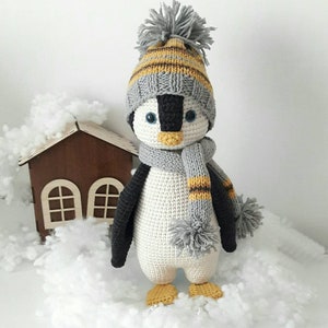 Könnte beinhalten: Ein gehäkeltes Pinguin-Spielzeug, das eine grau-gelb gestreifte Mütze mit Bommel und einen grau-gelb gestreiften Schal mit Bommeln trägt. Der Pinguin steht in einer verschneiten Szene.