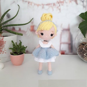 Könnte beinhalten: Eine gehäkelte Puppe mit blonden Haaren, einem blauen Kleid und weißen Schuhen. Die Puppe trägt eine Halskette und hat einen süßen Gesichtsausdruck.