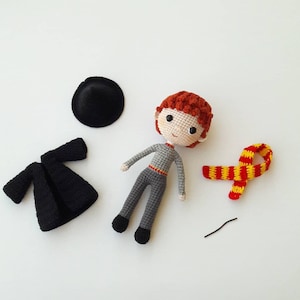 Könnte beinhalten: Häkelpuppe Amigurumi eines Jungen mit roten Haaren, der einen grauen Pullover und eine schwarze Hose trägt. Die Puppe hält einen rot-gelben Schal und einen schwarzen Hut.