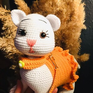 Könnte beinhalten: Weißes gehäkeltes Katzenspielzeug mit orangefarbener Häkelkleidung und einem gelben Knopf. Die Katze hat schwarze Augen und eine rosa Nase.