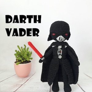 May include: A black crocheted Darth Vader plush toy holding a red lightsaber. The toy is standing on a white wooden surface with a small potted plant in the background. The text "DARTH VADER" is written in black letters above the toy.