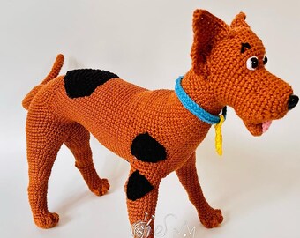 Puede incluir: Un juguete de peluche tejido a crochet de Scooby Doo, un gran danés, con pelaje marrón y manchas negras. El juguete está de pie sobre un fondo blanco y tiene un collar azul con una etiqueta amarilla.