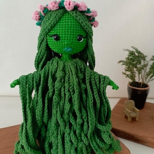 Puede incluir: Mu&ntilde;eca de crochet verde con el pelo largo y una corona de flores rosas. La mu&ntilde;eca tiene una cara verde con ojos negros y una boca rosa. La mu&ntilde;eca lleva un vestido de crochet verde.