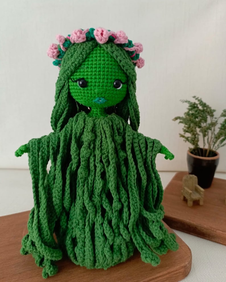 Puede incluir: Mu&ntilde;eca de crochet verde con el pelo largo y una corona de flores rosas. La mu&ntilde;eca tiene una cara verde con ojos negros y una boca rosa. La mu&ntilde;eca lleva un vestido de crochet verde.