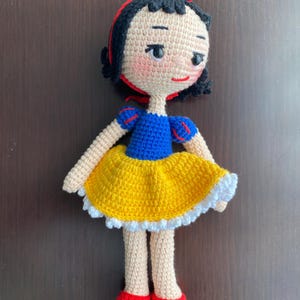 Könnte beinhalten: Eine gehäkelte Puppe von Schneewittchen, die ein gelbes Kleid mit einer weißen Spitzenborte, ein blaues Mieder und ein rotes Stirnband trägt. Die Puppe hat schwarze Haare und ein bemaltes Gesicht.