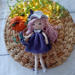 Könnte beinhalten: Eine gehäkelte Puppe mit einem lila Kleid, rosa Haaren und einem weißen Hut, die eine grüne Blume hält. Die Puppe sitzt auf einer geflochtenen Strohhutmatte mit einem kleinen orangefarbenen Kürbis.