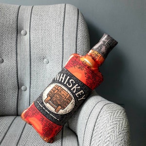 Op de afbeelding: Een decoratief kussen in de vorm van een whiskyfles met het woord "Whiskey" erop gedrukt. Het kussen is rood, oranje en zwart met een witte achtergrond.