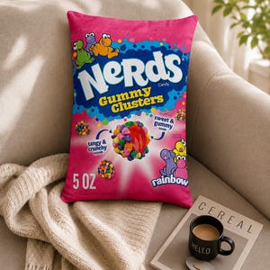 May include: A rectangular throw pillow featuring a vibrant pink design that mimics a bag of Nerds Gummy Clusters candy. The front displays the Nerds logo, colourful cartoon characters, a central gummy cluster graphic, and the text 5 OZ, rainbow, tangy & crunchy outside, and sweet & gummy inside.