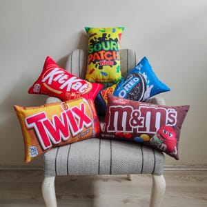 May include: A selection of candy-themed cushions, featuring brands like Kit Kat, Twix, M&M's, Oreo, and Sour Patch Kids. The cushions are in various shapes and sizes, with bright colours and striking text. They are arranged on a striped armchair.