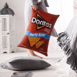 Könnte beinhalten: Ein rotes Kissen in Form einer Tüte Doritos Nacho Cheese Tortilla Chips. Das Kissen hat das Doritos-Logo und den Schriftzug "Party Size!". Eine Person in einem grauen Bademantel hält das Kissen.
