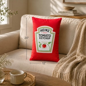 May include: A rectangular red pillow with the Heinz Tomato Ketchup label, measuring 20 inches long and 12 inches wide. The pillow is on a beige sofa with a beige throw blanket. A coffee table with a cup and vase is in the foreground.