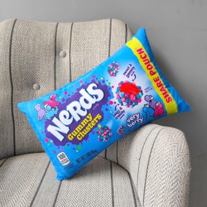 May include: A blue and yellow pillow shaped like a Nerds candy package. The pillow has the words "Nerds Gummy Clusters" and "Share Pouch" printed on it. The pillow is sitting on a gray and white striped chair.