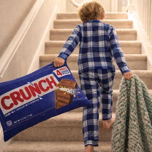 Puede incluir: Una persona en pijama a cuadros azules sube una escalera, llevando una gran almohada azul con forma de barra de chocolate Crunch y una manta verde y blanca. La almohada muestra el logotipo de Crunch y los detalles del producto.