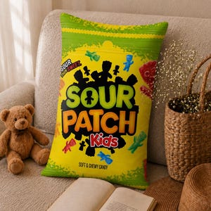 Könnte beinhalten: Ein rechteckiges Dekokissen mit dem Design der Sour Patch Kids Süßigkeitenverpackung. Das Kissen ist leuchtend gelb und grün und auf der Vorderseite mit dem Text SOUR PATCH Kids, SOUR THEN SWEET und SOFT & CHEWY CANDY sowie bunten Bonbonfiguren bedruckt.