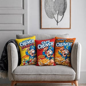 Może przedstawiać: Trzy prostokątne poduszki dekoracyjne z nadrukami opakowań płatków śniadaniowych Cap'n Crunch. Żółta poduszka przedstawia Crunch Berries, czerwona oryginalne Cap'n Crunch, a pomarańczowa Peanut Butter Crunch, każda z wizerunkiem maskotki.