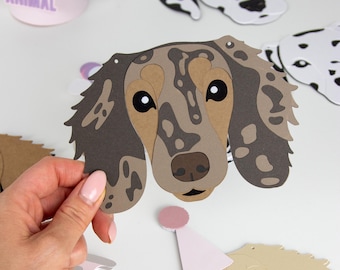 May include: A collection of dog-themed party decorations. The central piece is a paper cutout of a dachshund's face, featuring brown, tan, and black spots. Other dog faces, including a Dalmatian and a golden retriever, are also visible. A party hat with the words "PARTY ANIMAL" is in the background.
