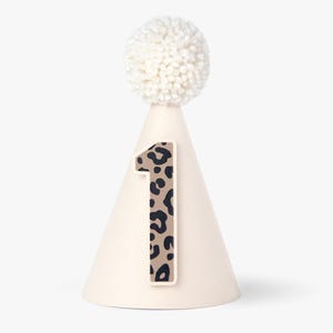 Könnte beinhalten: Weißer, kegelförmiger Partyhut mit einem weißen Bommel oben. Der Hut hat eine braune und schwarze Leopardenmuster-Eins auf der Vorderseite.