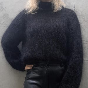 May include: A black, fluffy knit sweater with a high neckline and long sleeves. The sweater is paired with black leather pants. The person is leaning against a gray wall.