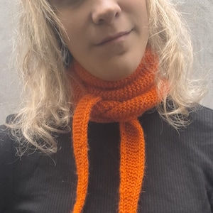 May include: A bright orange knitted scarf with long, hanging ends. The scarf is a vibrant orange colour and looks handmade. The person in the image is wearing a black long-sleeved top.