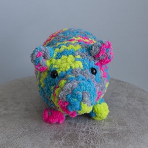 May include: A colourful crocheted stuffed animal shaped like a pig. The pig is made with grey, blue, pink, and yellow yarn. It has black eyes and a pink nose.
