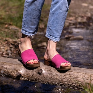 May include: A pair of pink suede clogs with wooden soles. The clogs are worn by a person standing on a wooden log over a stream.