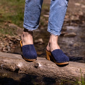 Puede incluir: Un par de zuecos de gamuza azul con tacones de madera y un borde tachonado. Los zuecos los lleva una persona de pie sobre un tronco de madera.